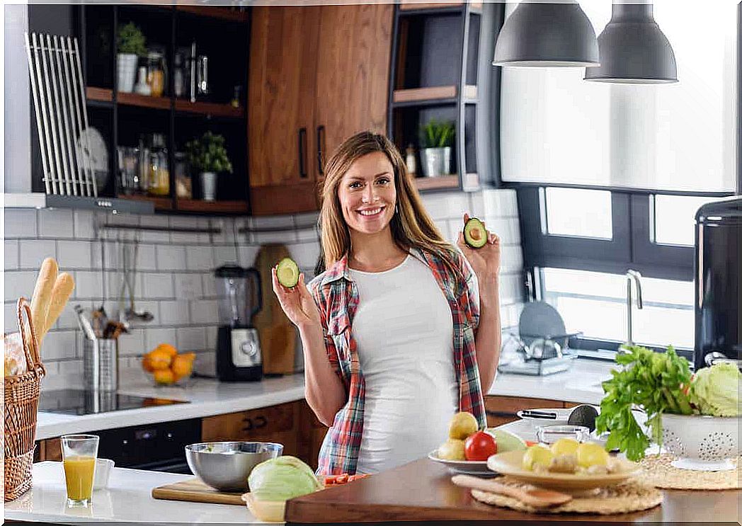 A pregnant woman in a kitchen holding two halves of an avocado and smiling.