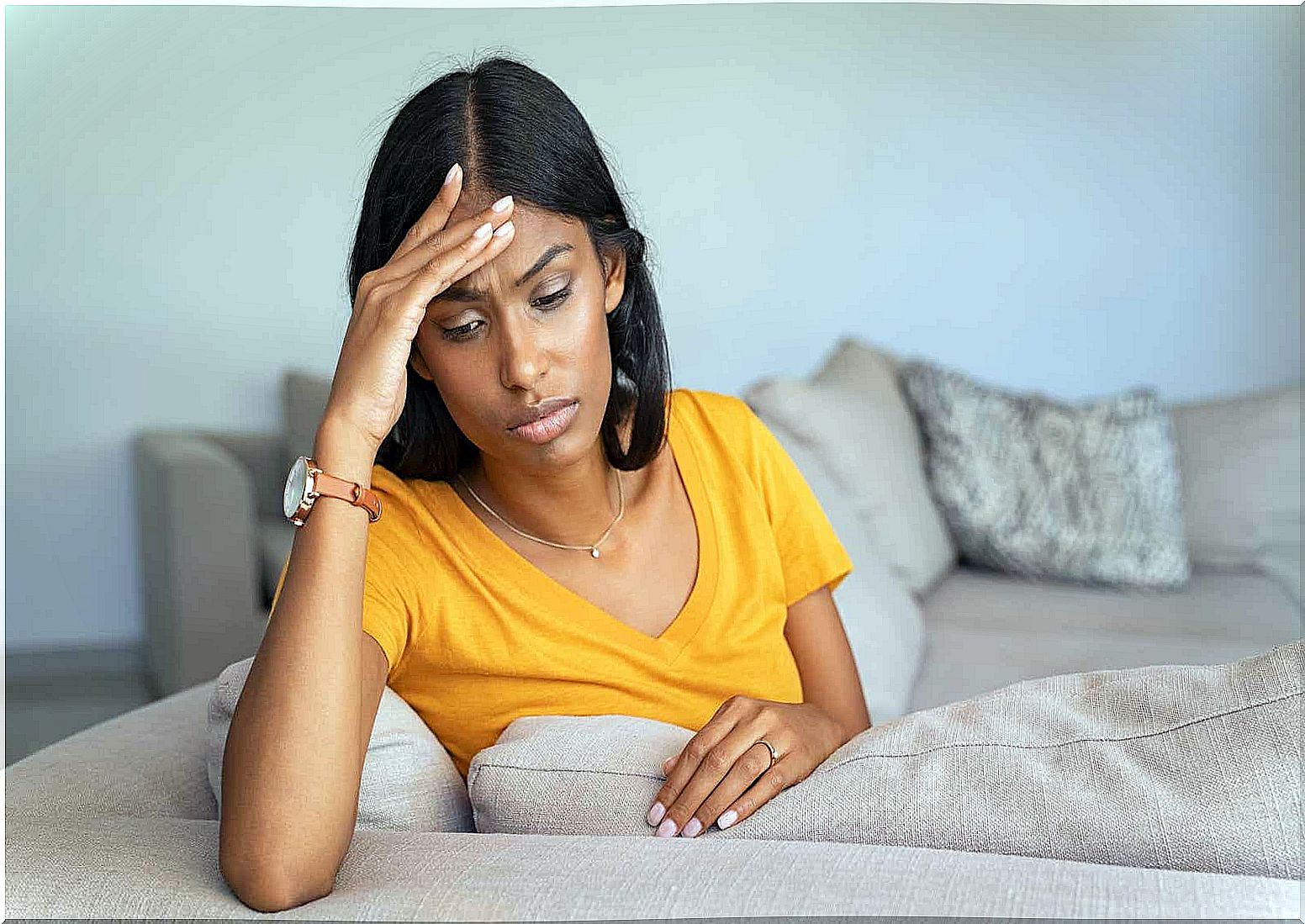 A woman sits on a sofa with her hand on her forehead, frowning.